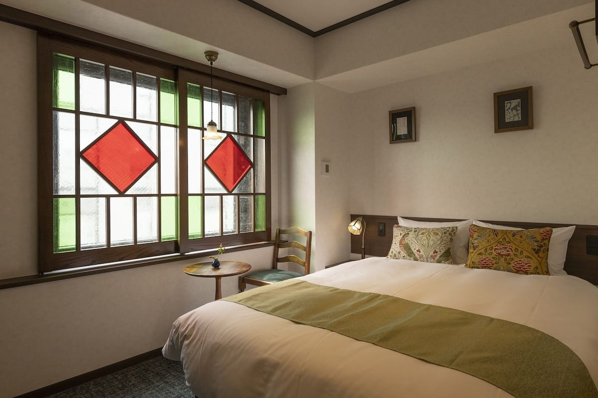 Habitación de hotel con una cama grande, una mesita pequeña, una silla y un ventanal con vidrieras.