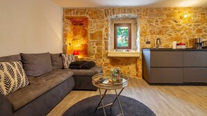 A living room with a stone wall, a sofa, a round coffee table, and a kitchen area. - Apartment Kleine Perle Am Meer (Silo - island Krk)