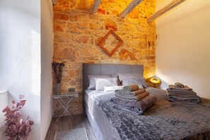A bedroom with a stone wall, a bed with a grey headboard, and a bedside table with a lamp.