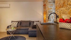 A modern living room with a sofa, a small round table, and a kitchen area with a stone wall and sink. - Apartment Kleine Perle Am Meer (Silo - island Krk)