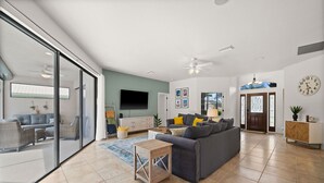 A spacious living room with a grey sofa, a wooden side table, and a large window. - Family Friendly 3BR Beach House with salt water heated pool (Englewood)