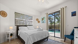 A bedroom with a bed, a nightstand, a chair, a large window with blinds, and a view of a pool. - Family Friendly 3BR Beach House with salt water heated pool (Englewood)