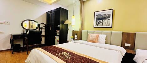 A hotel room with a large bed, a desk with a chair, a wardrobe, a lamp, and a framed picture on the wall.