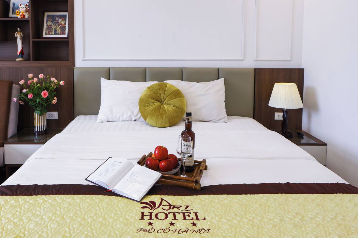 Habitación de hotel con una cama, una mesita de noche con lámpara, un florero con flores y una bandeja con manzanas y una botella.