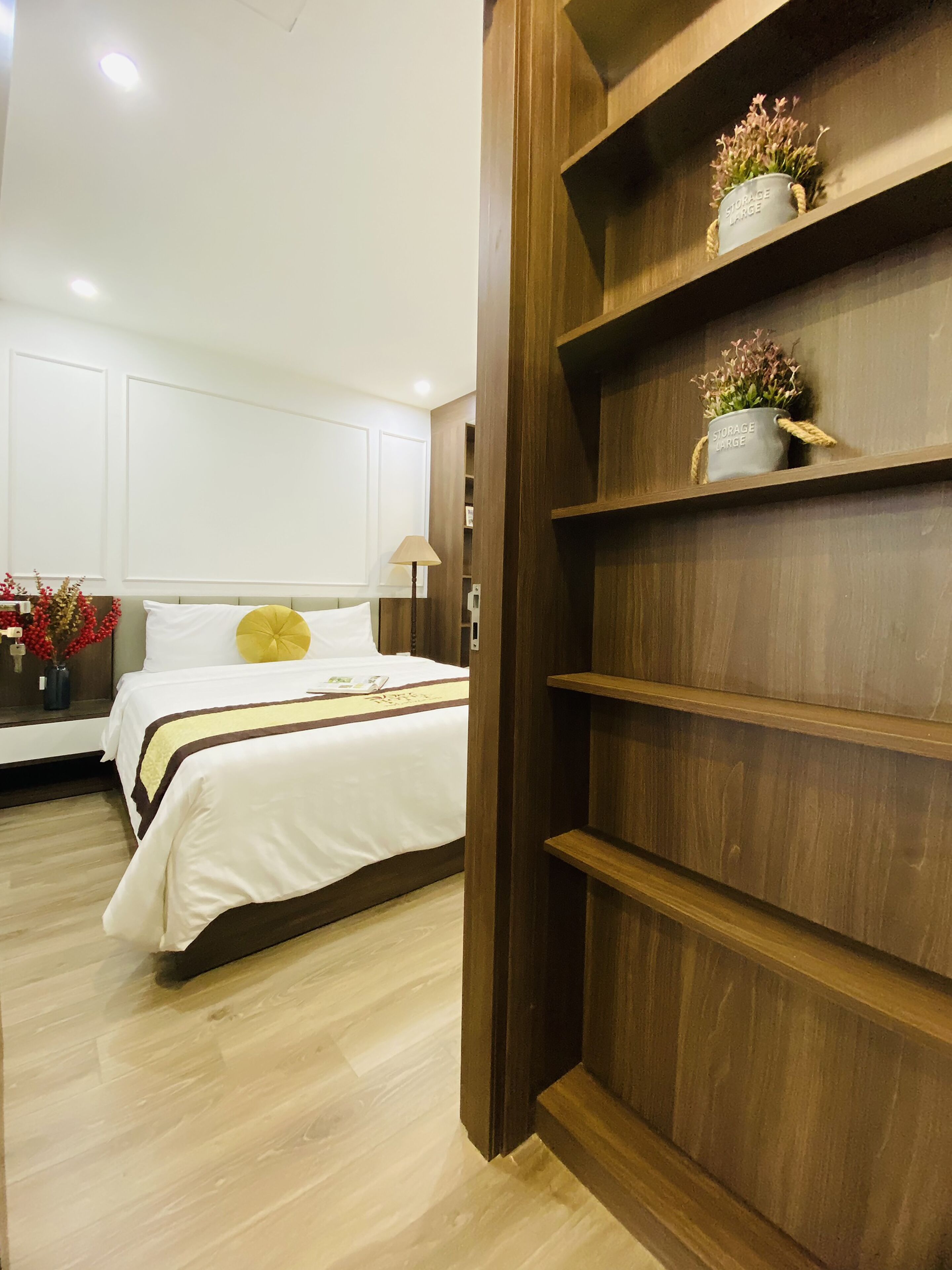 A hotel room with a bed, a wooden shelf with a vase, and a white wall.