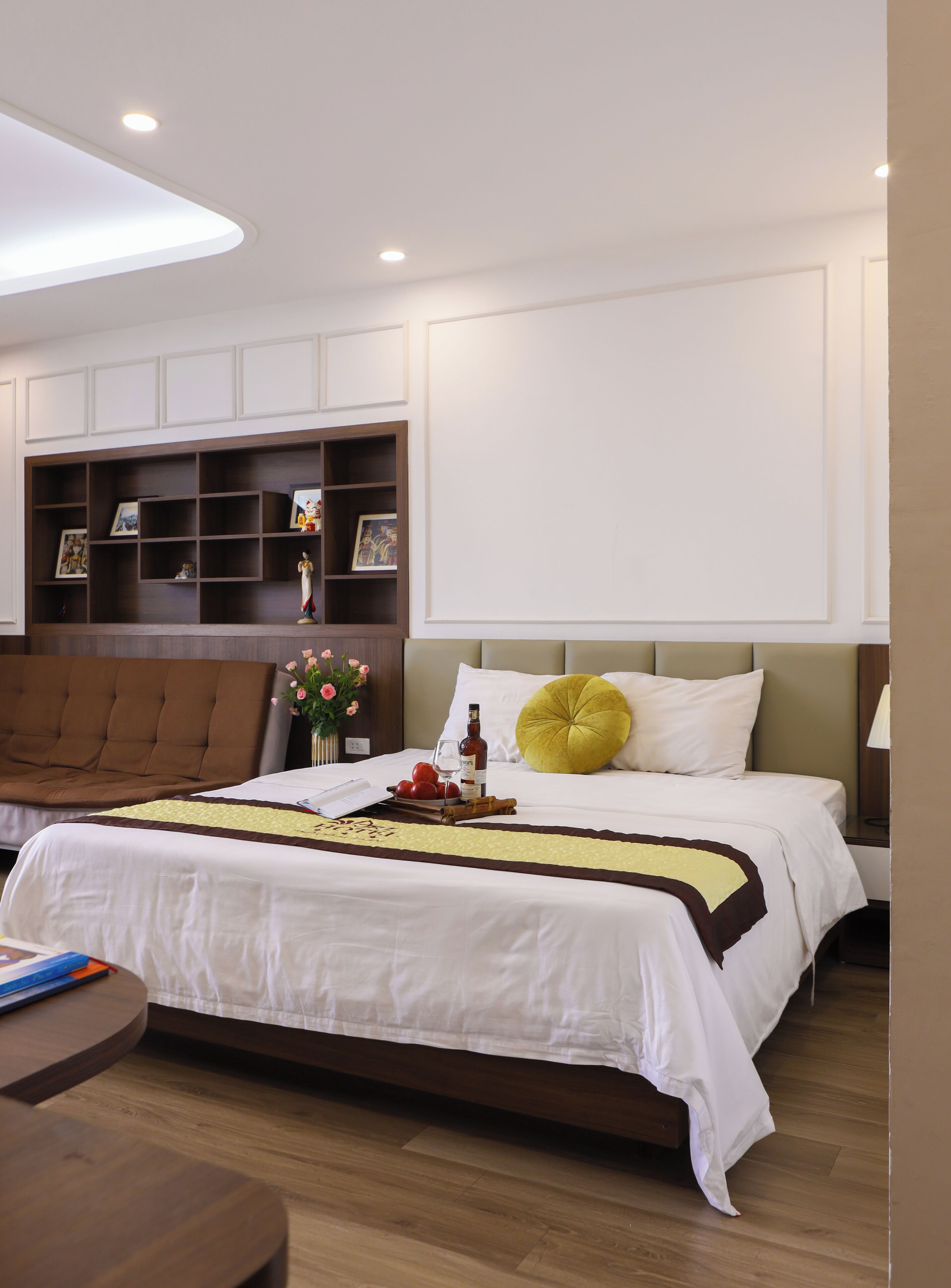 A neatly arranged bedroom with a large bed, a wooden headboard, a bedside table, and a built-in shelf with framed pictures.