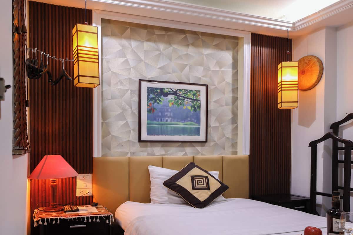 Una habitación de hotel con una cama, una mesita de noche, una lámpara, un cuadro en la pared y una botella sobre la mesita.