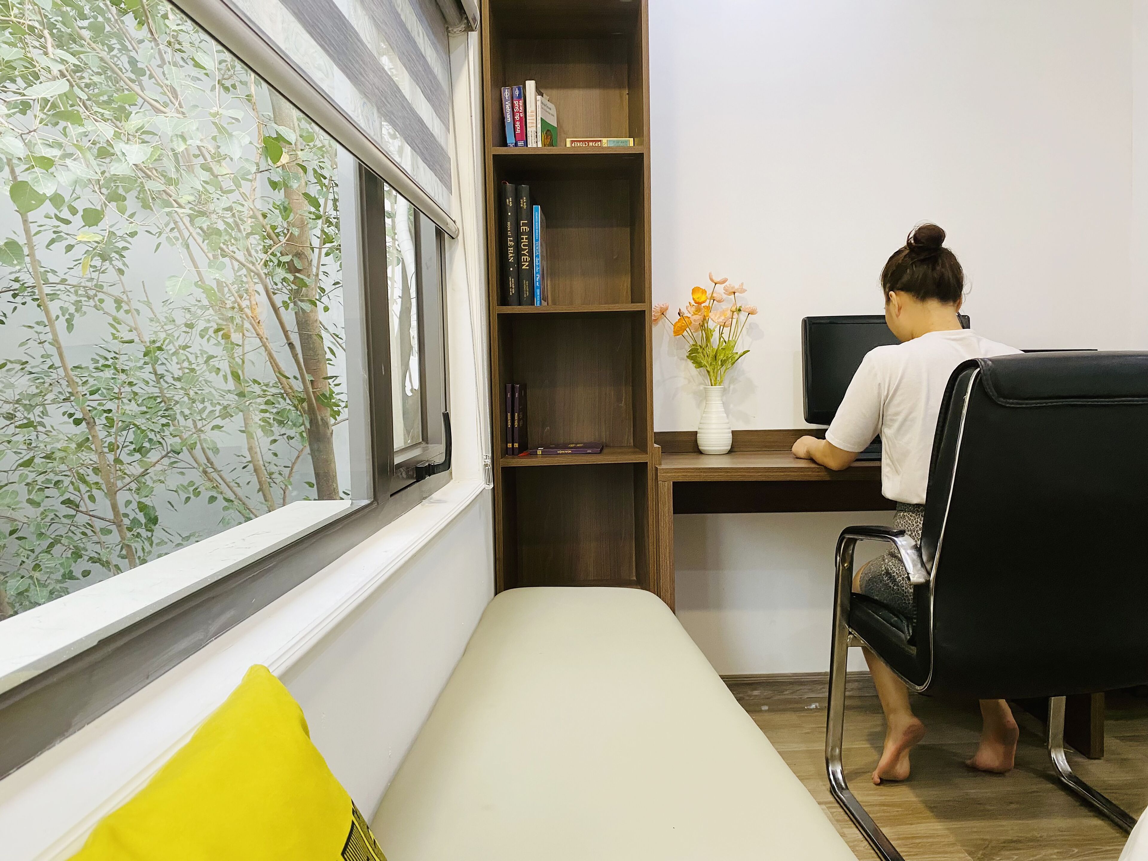 A room with a desk, a chair, a bookshelf, a window with a view of trees, and a bed with a yellow pillow.