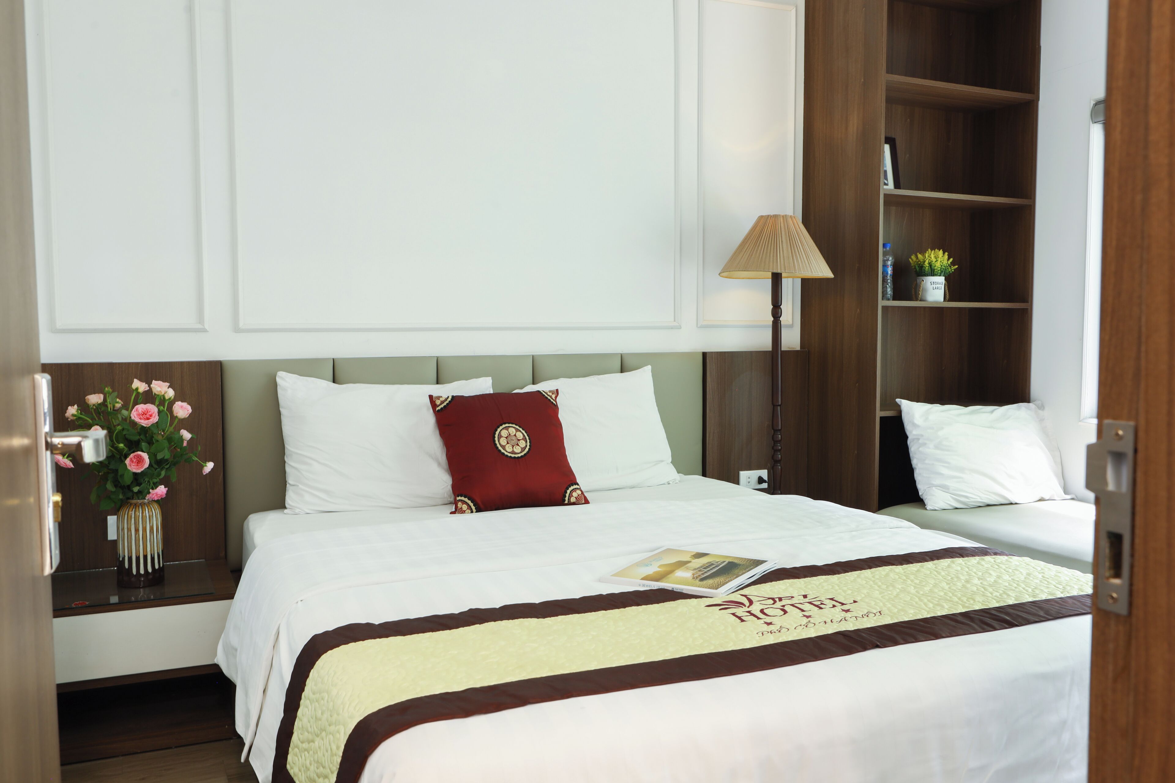 Una habitación de hotel con una cama, una mesita de noche con un jarrón de flores, una lámpara y una estantería.