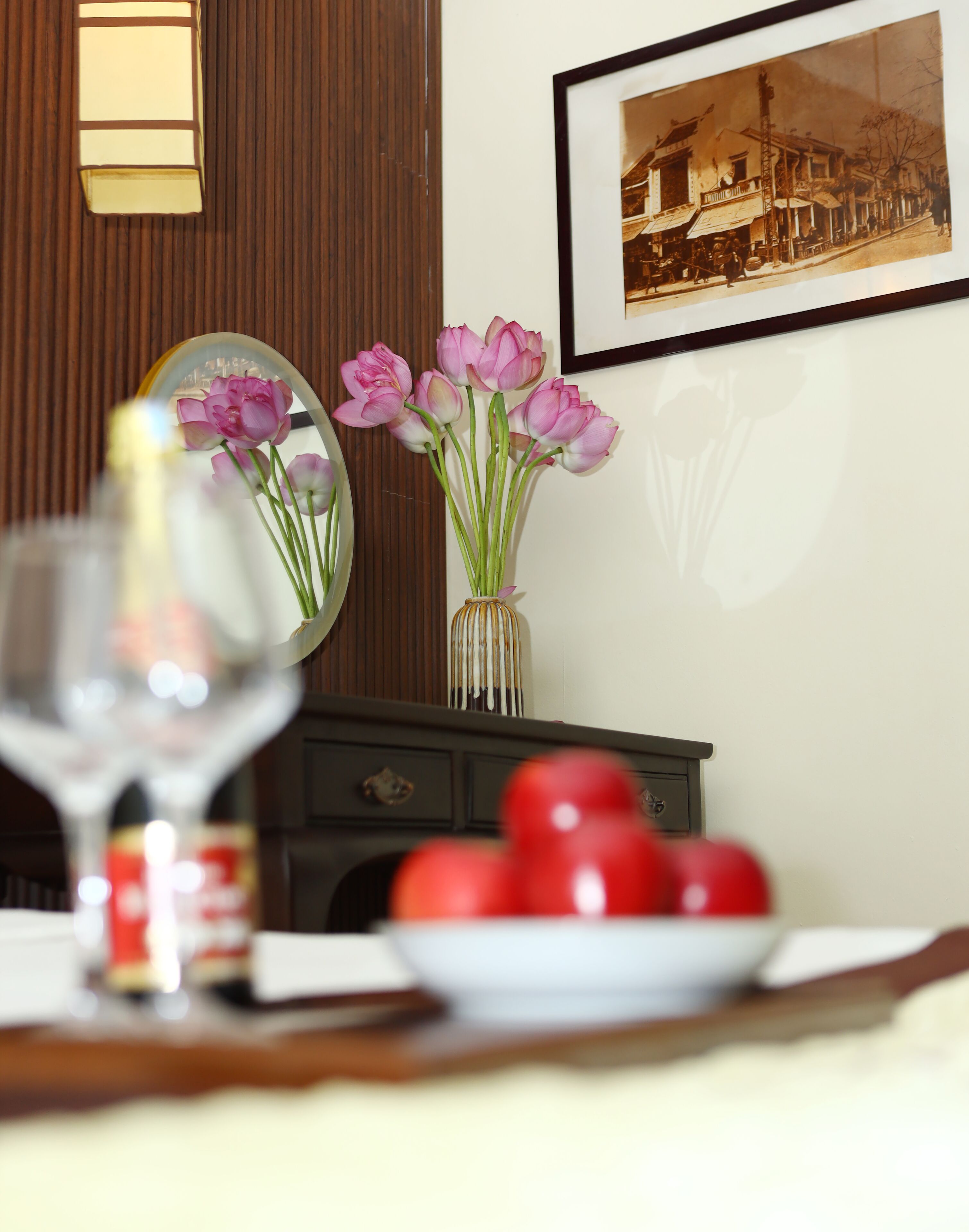 A dining table with a wine glass, a vase with pink flowers, and a plate of tomatoes.
