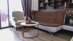 A living room with a brown sofa, a white armchair, a round coffee table with fruits and a bottle, and a bookshelf with decorative items. - Art Hotel Ha Noi (Hanoi)