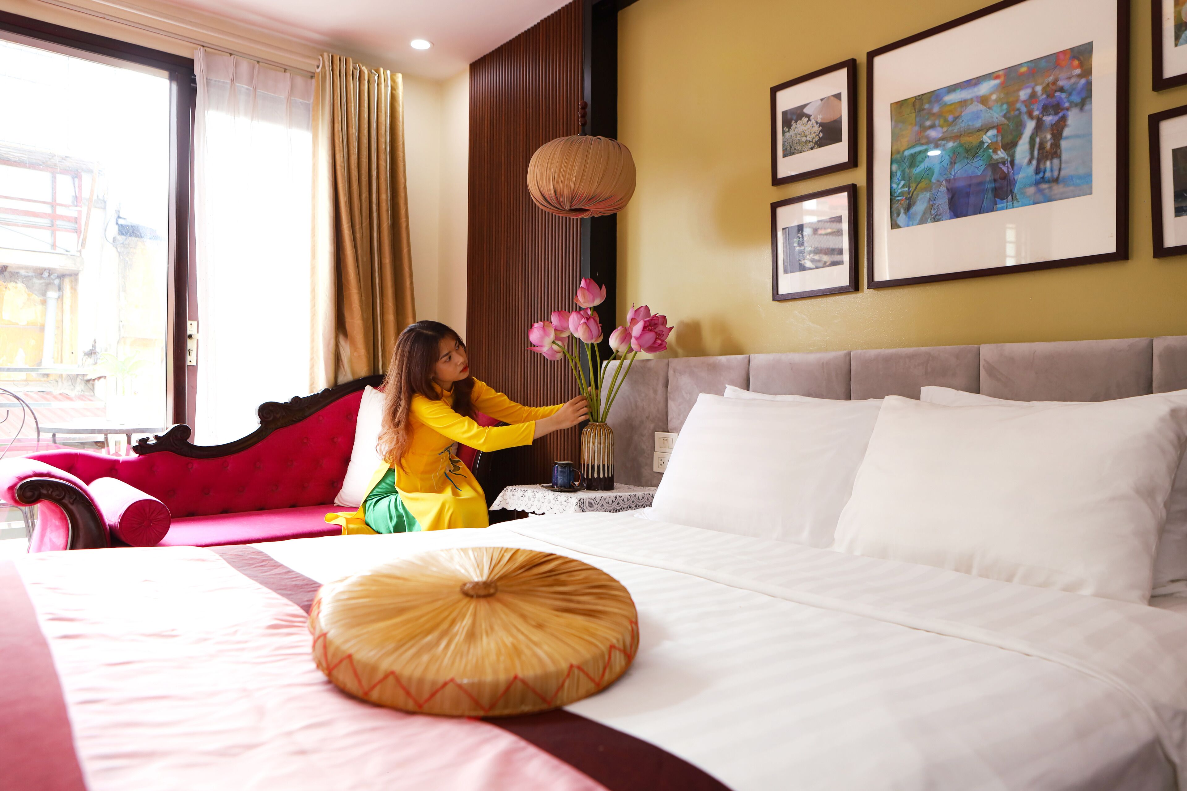 A hotel room with a bed, a red sofa, a vase of pink flowers, and framed pictures on the wall.