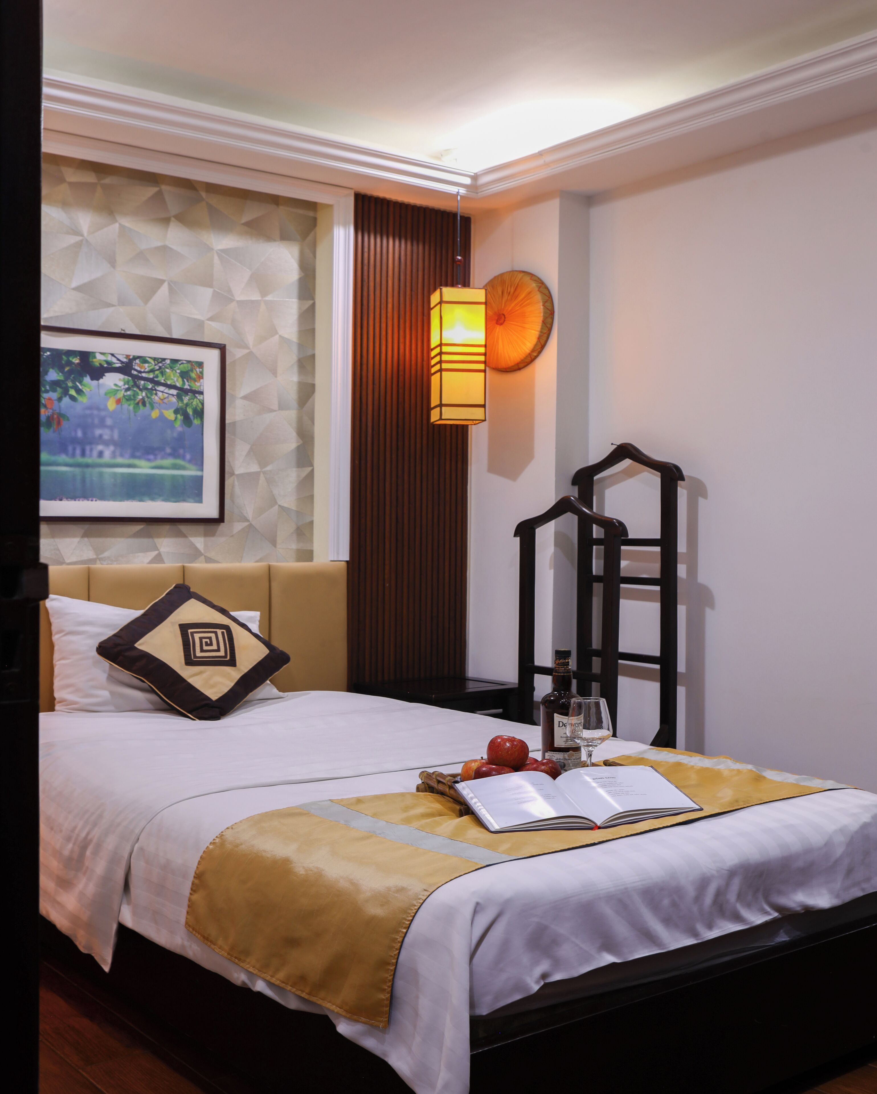 Uma cama bem arrumada com colcha branca e dourada, um quadro na parede, uma mesinha de cabeceira com garrafa e copo, e uma cadeira encostada à parede.