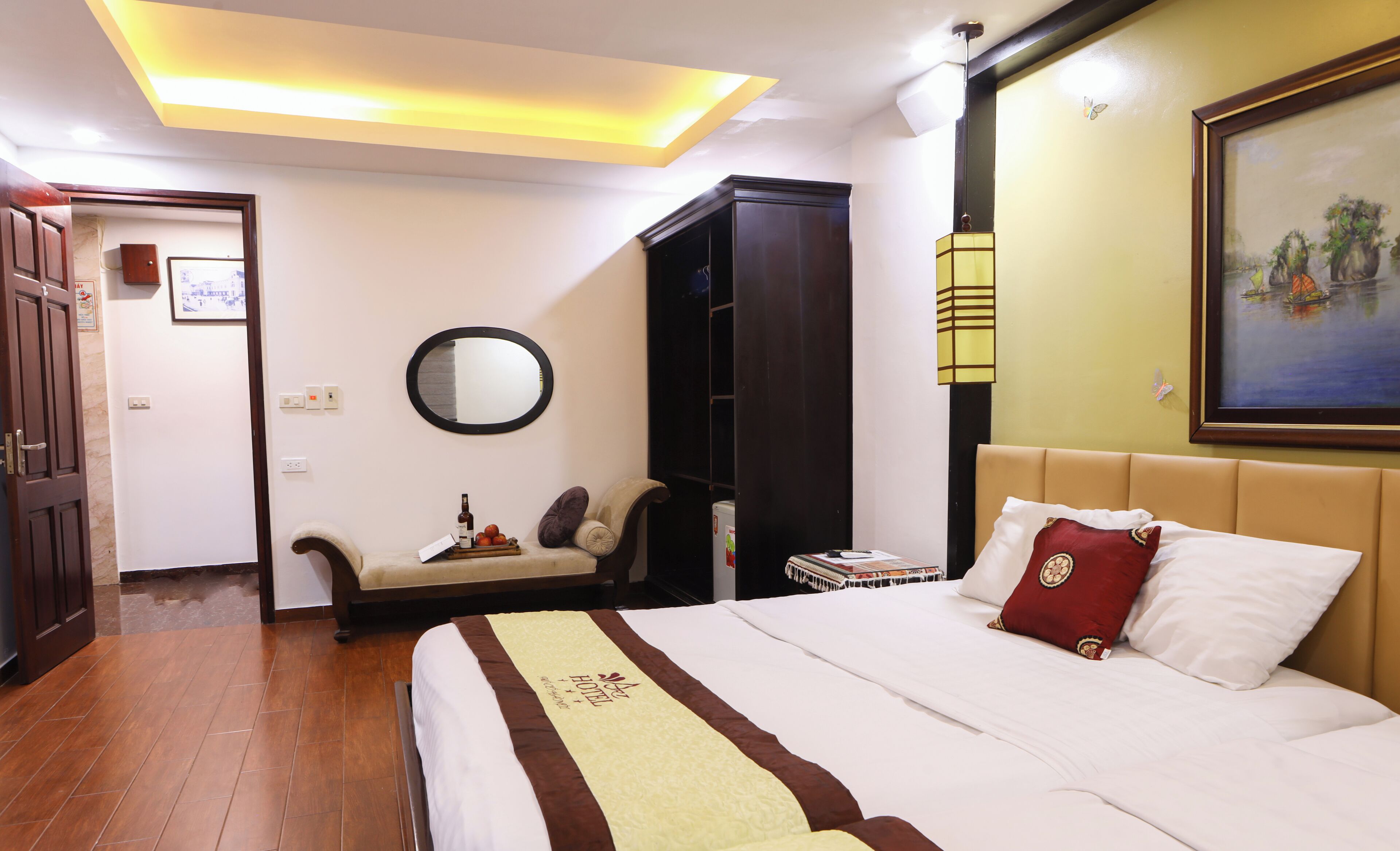Une chambre d’hôtel avec un lit, un miroir, une armoire, une table de chevet avec une bouteille et des fruits, et un tableau accroché au mur.
