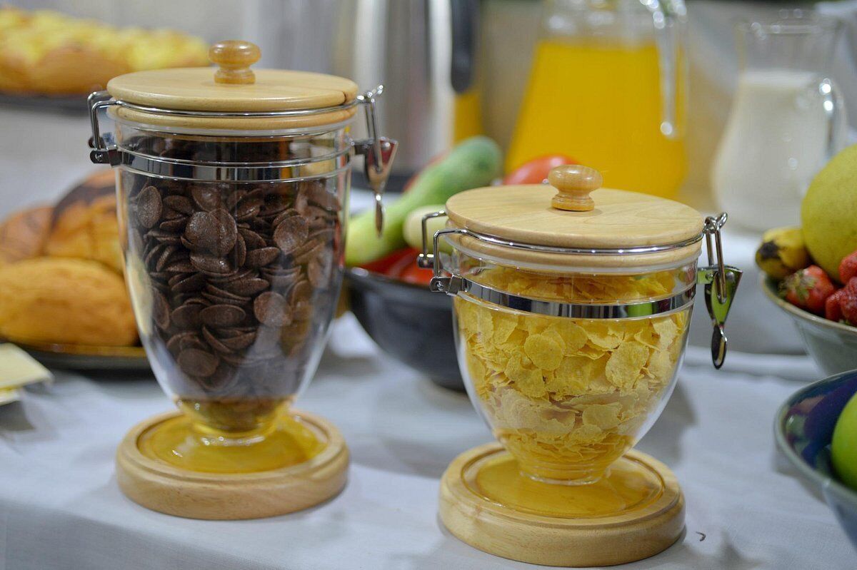 Twee glazen potten met houten deksels, gevuld met koffiebonen en ontbijtgranen, op een tafel met verschillende soorten fruit en een pitcher sinaasappelsap op de achtergrond.