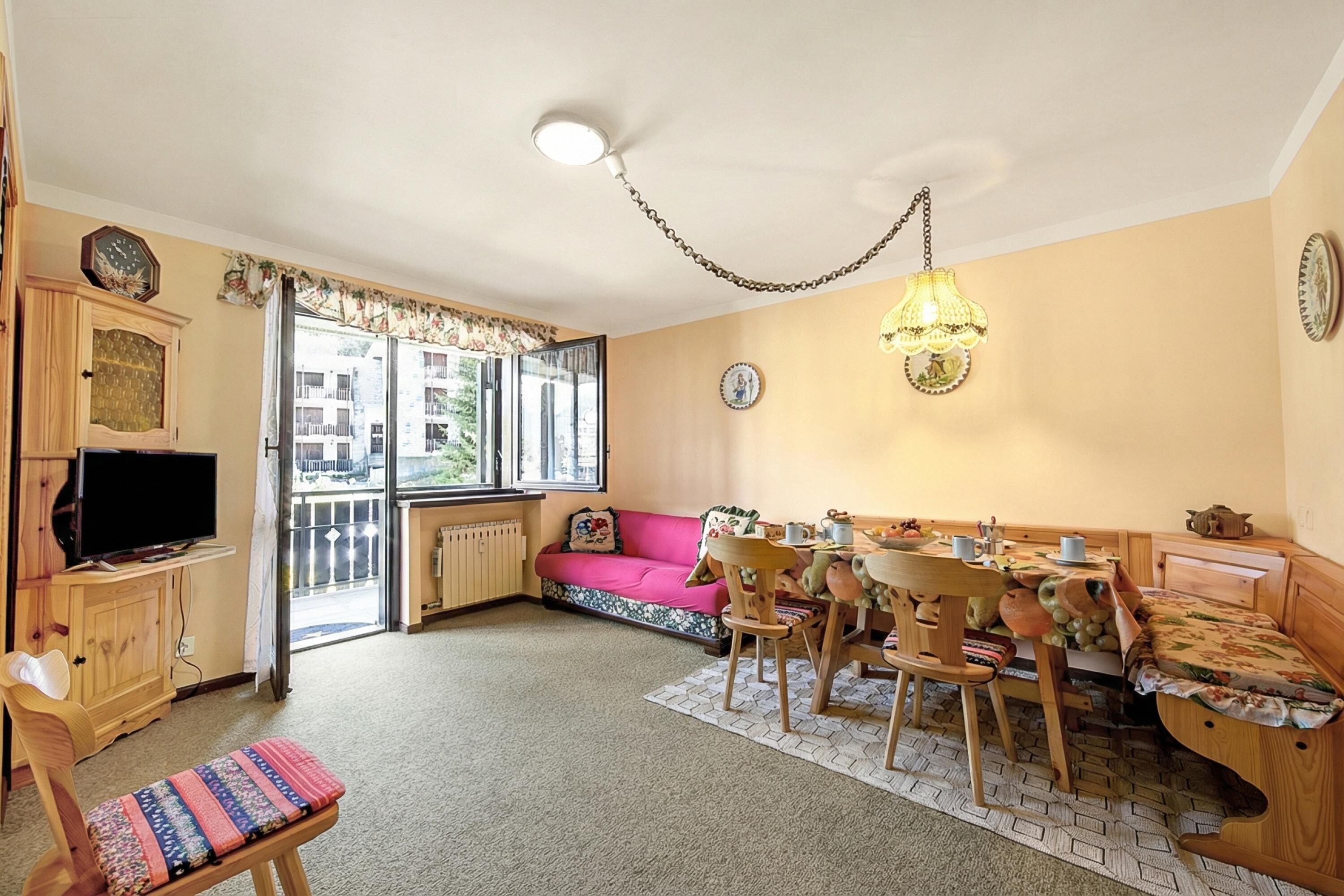 A cozy living room with a sofa, dining table, and chairs. There is a television on a wooden cabinet, and a window with a view of buildings outside.