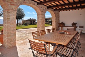 An outdoor patio with a wooden table and benches, brick arches, and a view of a house in the background.
