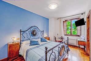 A bedroom with a bed, a nightstand, a desk, a window with a view, and a flat-screen TV mounted on the wall.