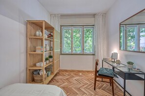A bedroom with a wooden bookshelf, a desk with a chair, a bed, and a window with curtains.