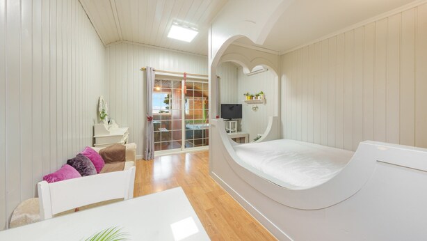 Una habitación con una cama curvada, una mesita pequeña, un escritorio blanco, un televisor y vistas a un área exterior con terraza de madera.