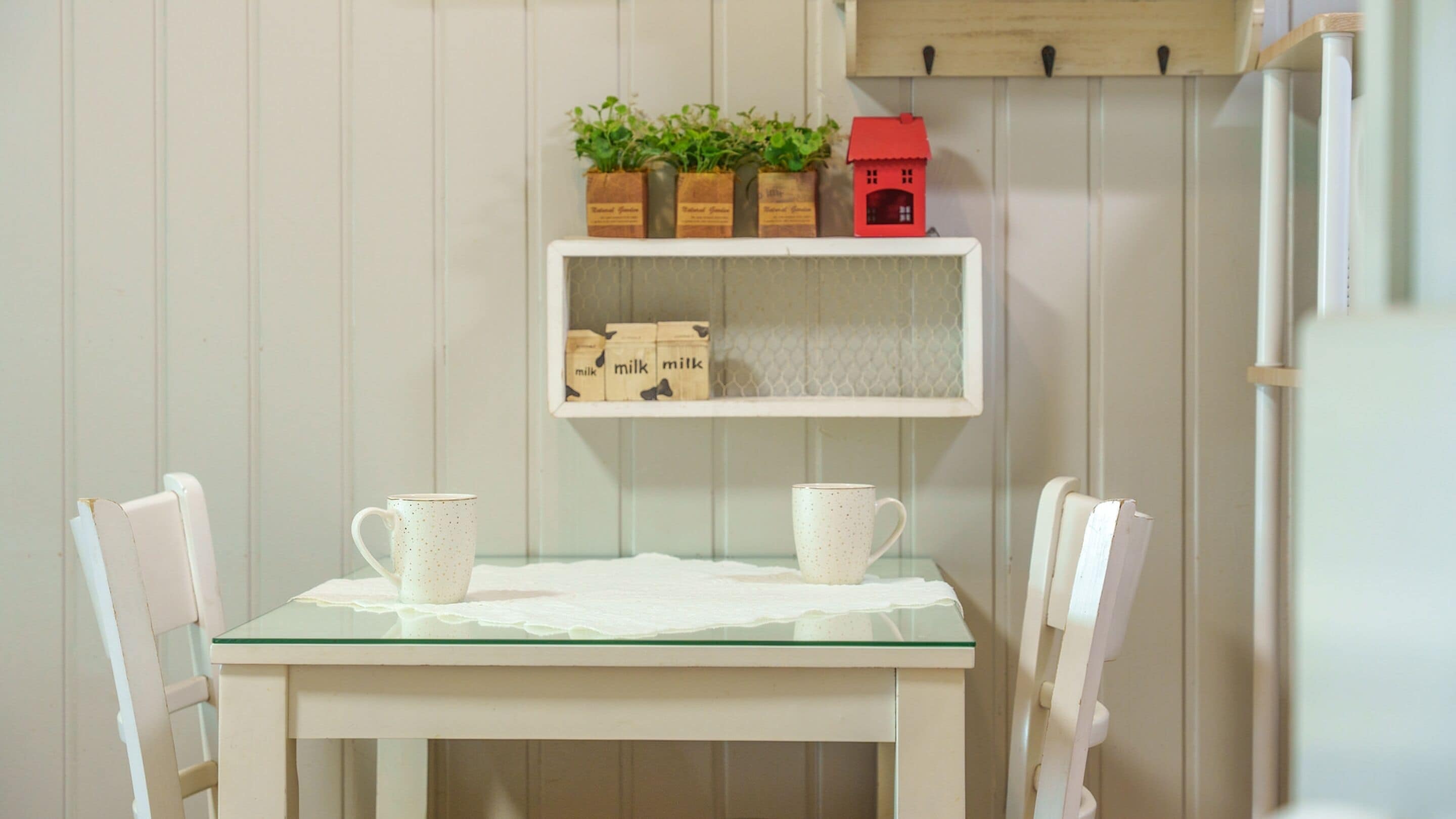 A small dining area with a glass-top table, two white chairs, and a shelf above holding potted plants and milk cartons.