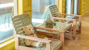 Un rincón acogedor con sillas y mesas de madera, un osito de peluche en una silla y una planta pequeña sobre una mesa.