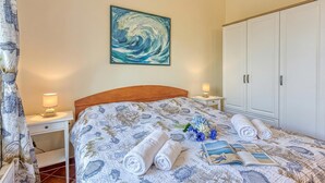 En pænt redt seng med et mønstret sengetæppe, et vægmaleri, et hvidt skab og et sengebord med en lampe.