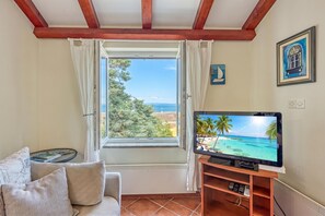 Ein Zimmer mit Fernseher, der eine Strandansicht zeigt, einem Fenster mit Vorhängen, einem kleinen Tisch mit Büchern und einem gerahmten Bild an der Wand.