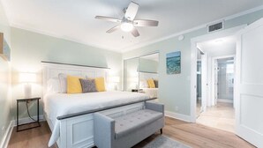 Une chambre à coucher comprenant un lit, des tables de chevet, un banc, un ventilateur de plafond et un grand miroir.