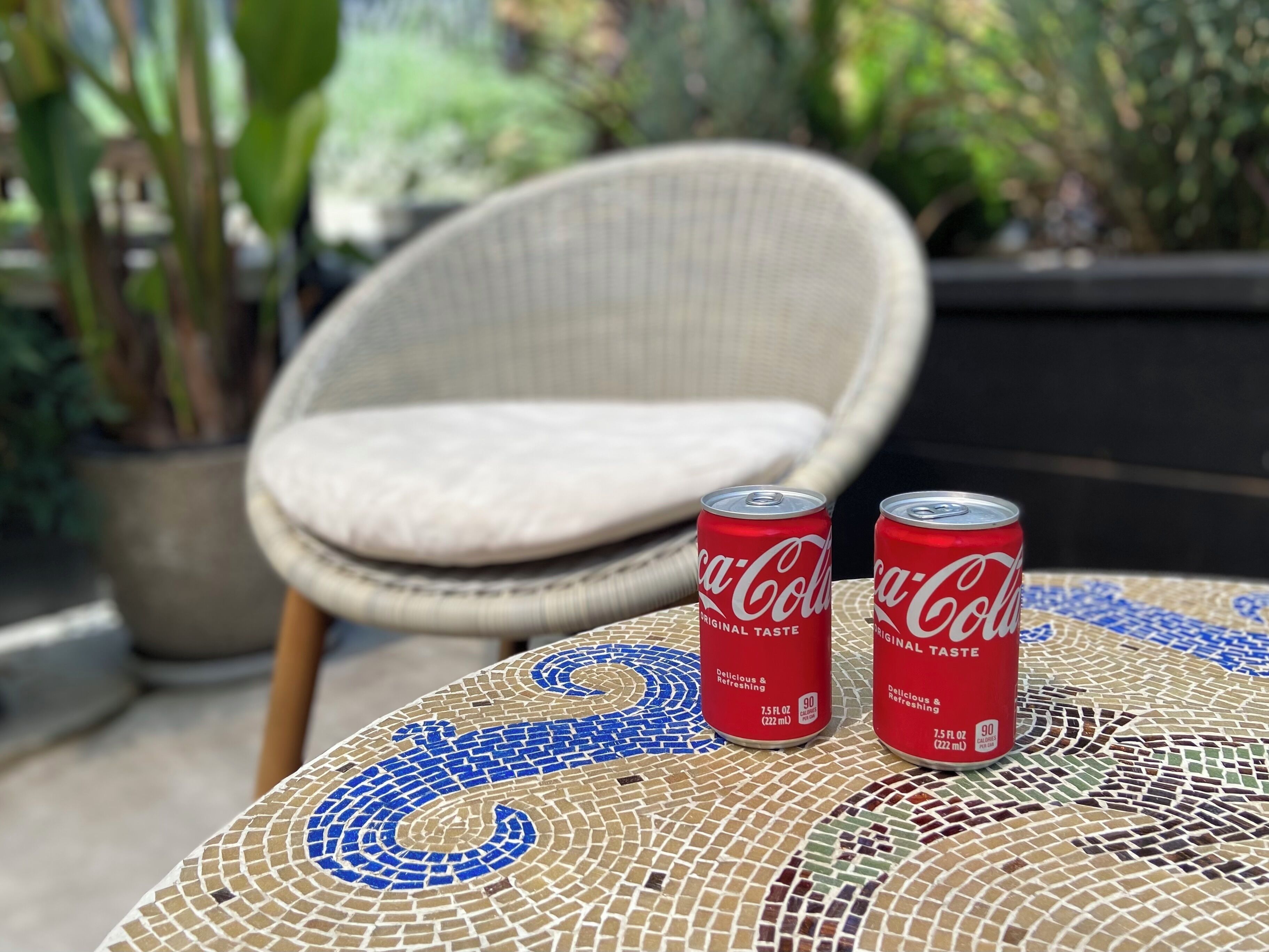 Deux canettes de Coca-Cola sur une table ornée d’un motif en mosaïque, avec une chaise en osier en arrière-plan.