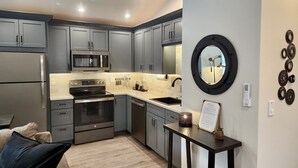 Une cuisine moderne avec des meubles gris, un réfrigérateur en acier inoxydable, un four à micro-ondes, un four et un évier. Un miroir rond est accroché au mur, et une petite table porte une bougie.