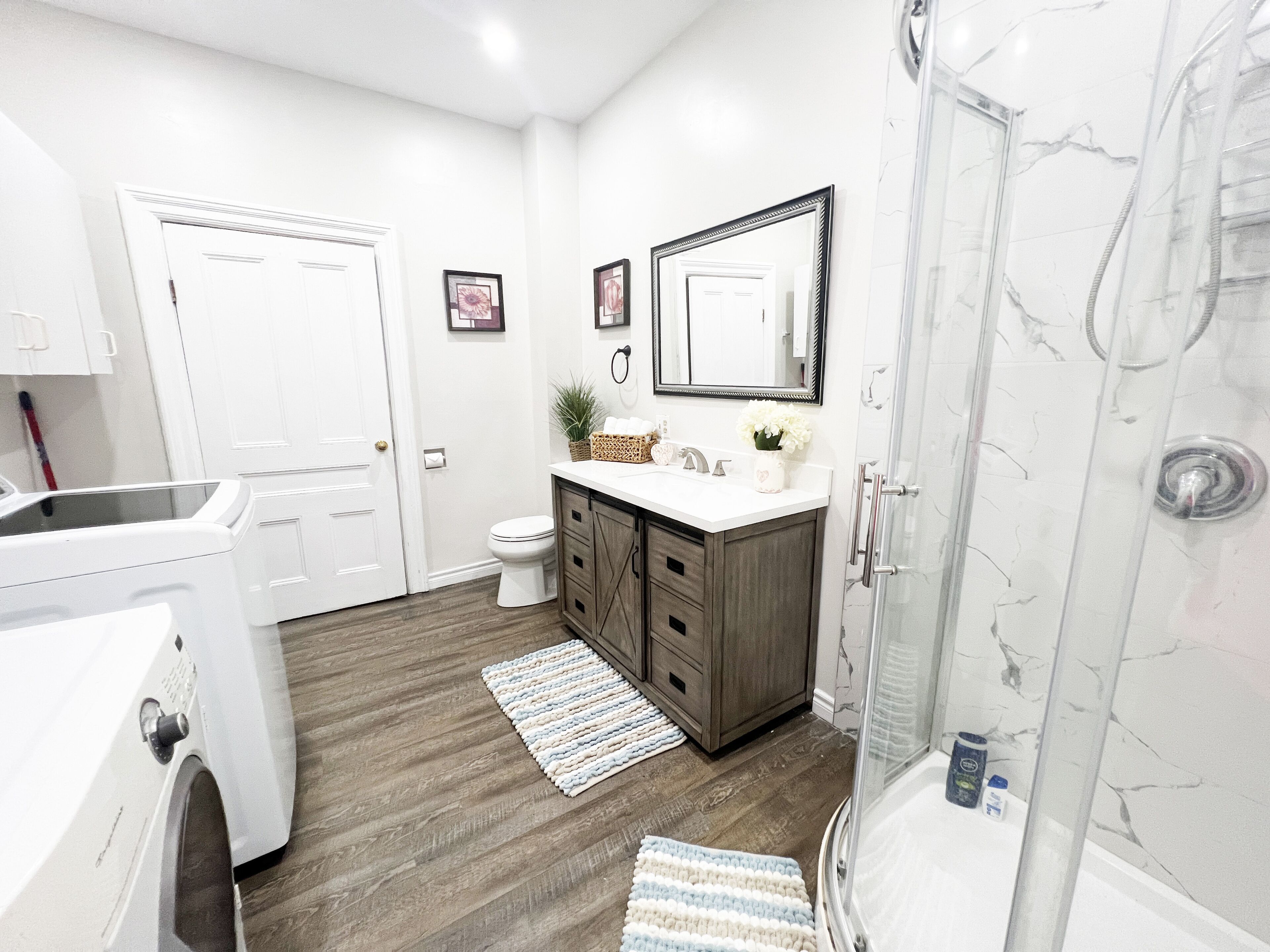 Une salle de bain moderne avec un lavabo blanc, des meubles en bois foncé, un grand miroir et une douche dotée d’une porte en verre.