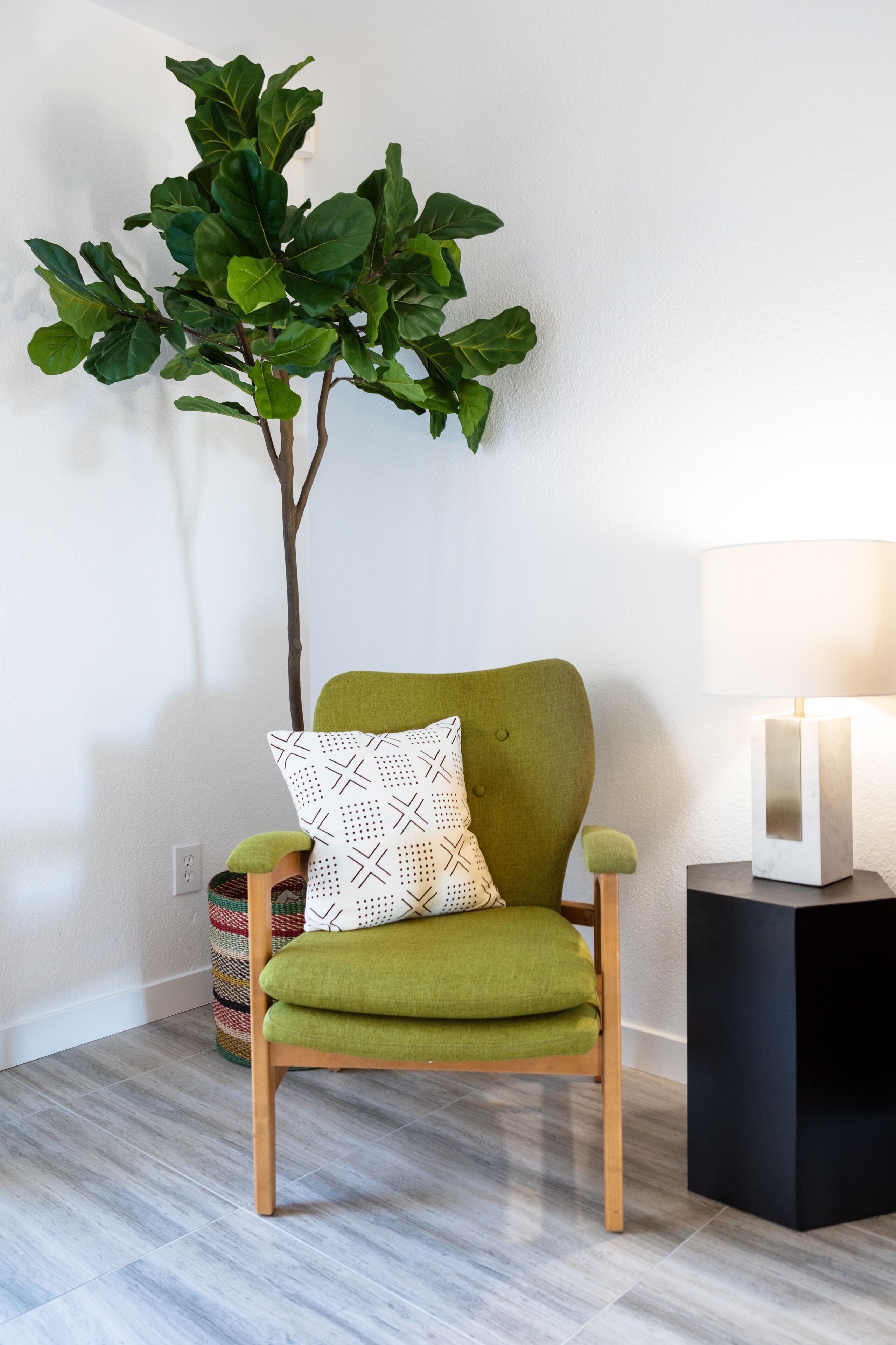 Een groene beklede stoel met een gestreept kussen, een bijzettafeltje met een lamp en een potplant voor een witte muur.