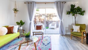 A bright living room with a sofa, armchair, and a coffee table. There is a large window with curtains, a potted plant, and a view of the outside.