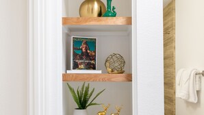 A narrow hallway with white walls and a wooden floor. Shelves are mounted on the wall, displaying various decorative items including a book, a green vase, and a golden spherical object.