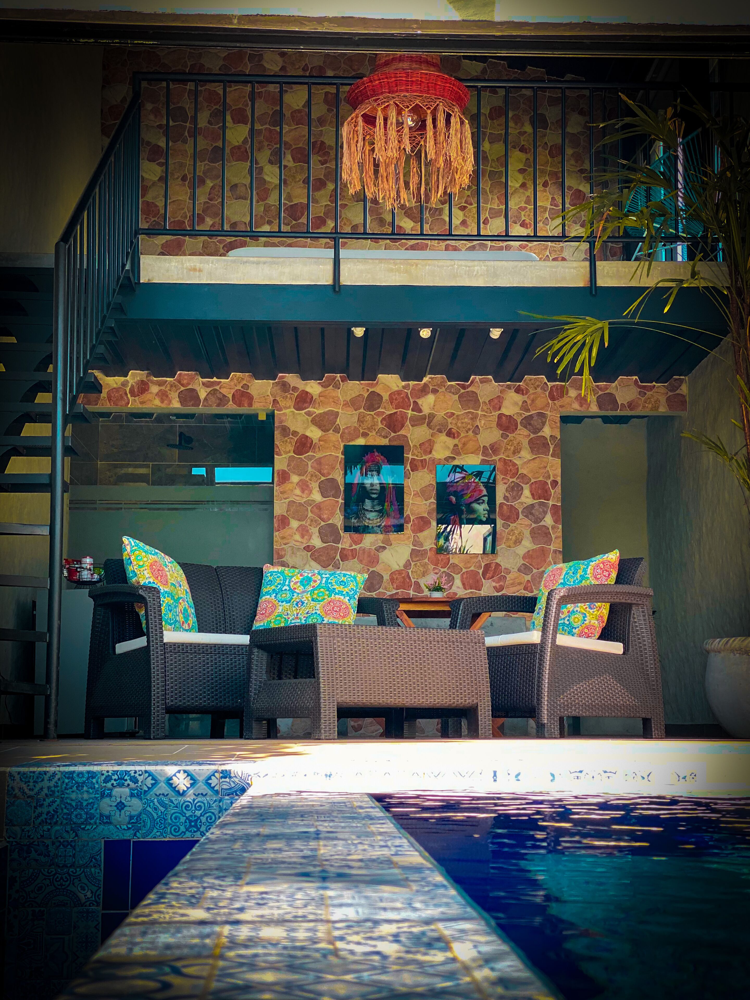 A two-story outdoor area with a stone wall, a balcony with a red hanging light, wicker furniture, and a pool.