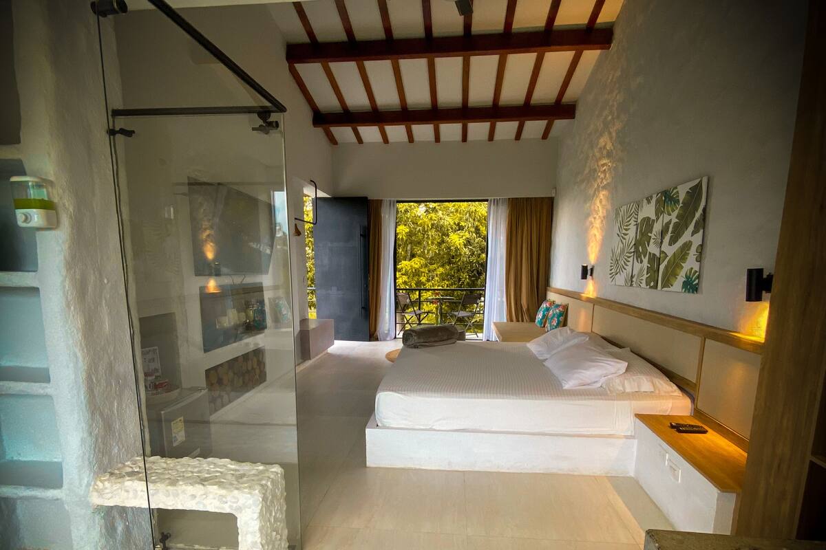 A modern bedroom with a glass shower, a bed with white linens, a wooden ceiling, and a view of greenery outside.