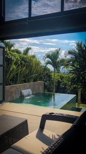 A modern outdoor pool area with a waterfall feature, surrounded by lush tropical plants and a clear blue sky. - ECOPARADISE (Pereira)