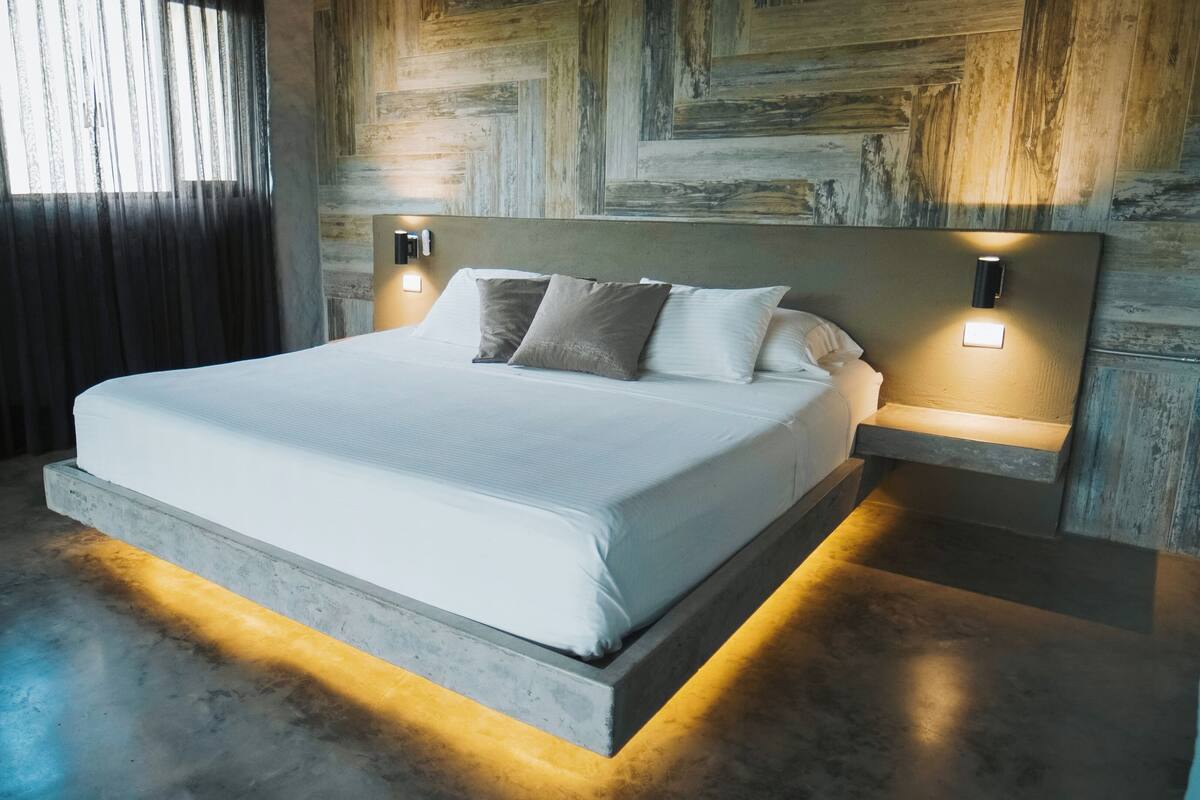 A modern bedroom with a large bed, two bedside tables with wall-mounted lights, a window with curtains, and a concrete platform under the bed.