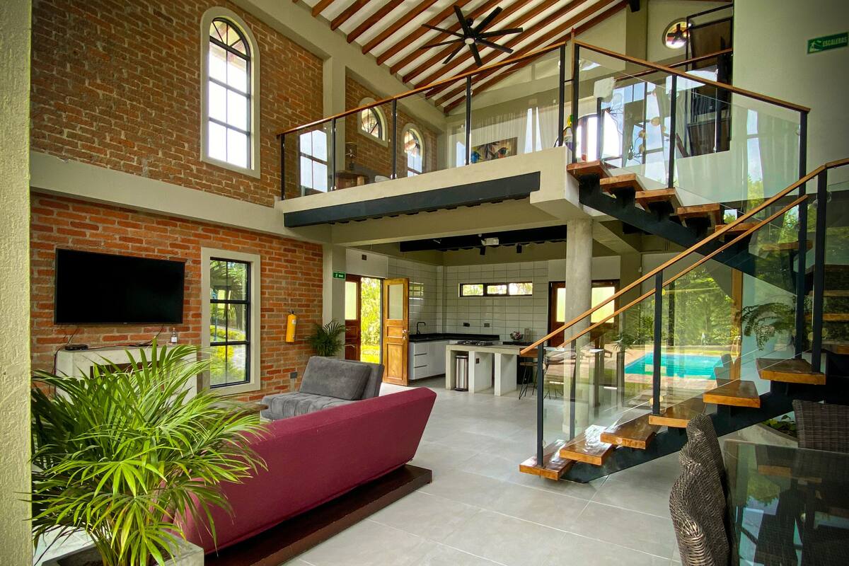 A spacious living area with a high ceiling, exposed brick walls, a modern sofa, and a glass staircase leading to an upper level.