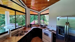 Une cuisine moderne équipée d’une cuisinière à gaz, d’un four et d’un réfrigérateur en acier inoxydable. De grandes fenêtres offrent une vue sur la verdure et les fleurs.
