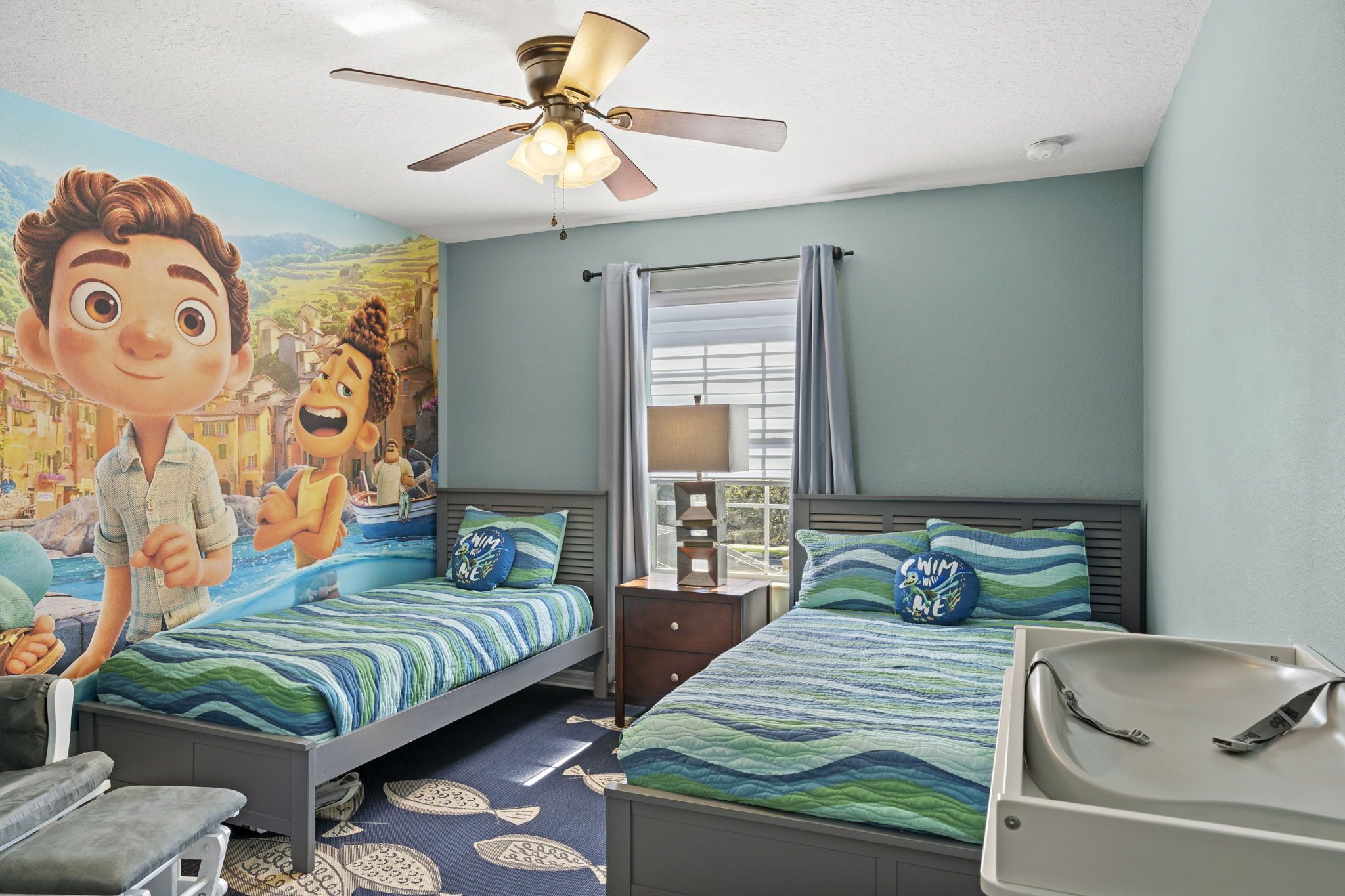 A bedroom with two beds, a ceiling fan, a window with curtains, and a mural on the wall.