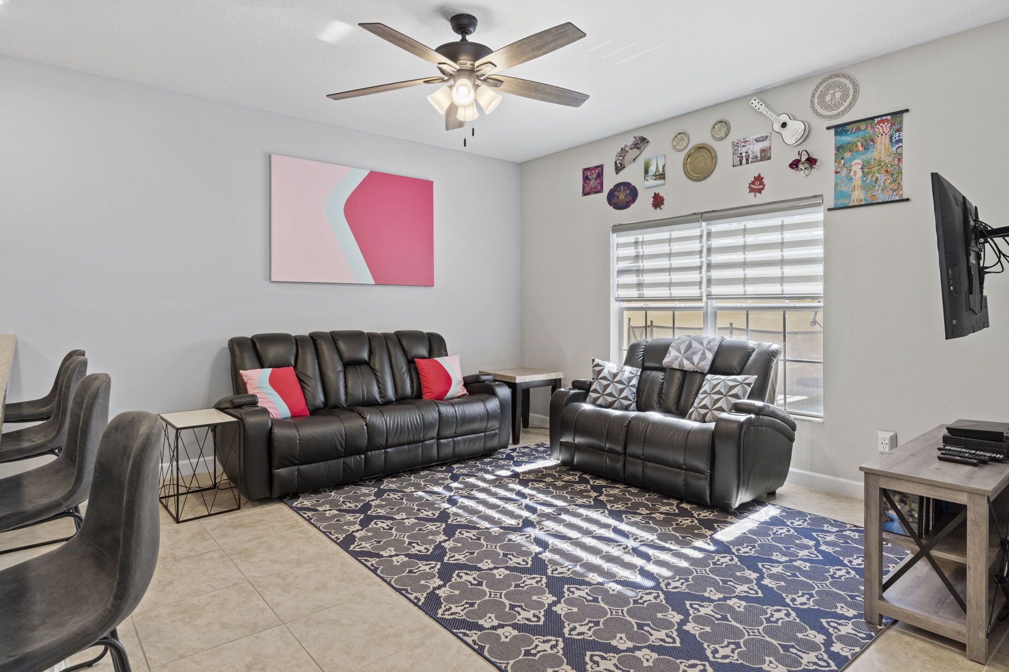 A living room with a ceiling fan, a large window with blinds, a black leather sofa, a coffee table, and a patterned rug.