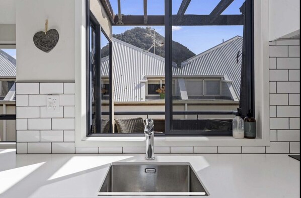 A modern kitchen with a stainless steel sink, a heart-shaped decoration on the wall, and a view of a house with a metal roof through a large window.