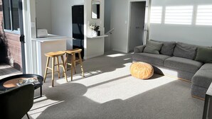 Een moderne woonkamer met een grijze bank, een rond voetenbankje en een salontafel met tijdschriften. Er is een keukenhoek met een koelkast en een eettafel met krukken.