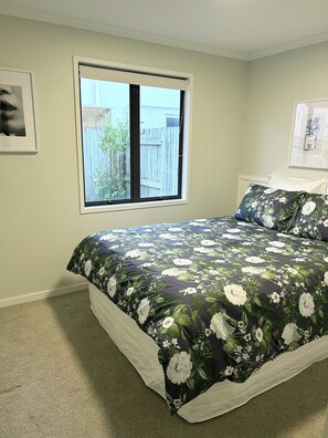 Une chambre avec une grande fenêtre, un lit recouvert d’une literie à motifs floraux et un tableau encadré accroché au mur.