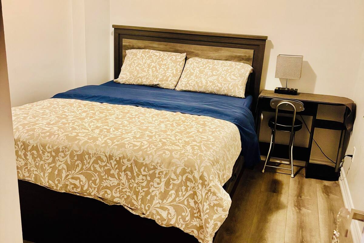 A neatly made bed with a patterned bedspread and pillows, a small bedside table with a lamp, and a chair.