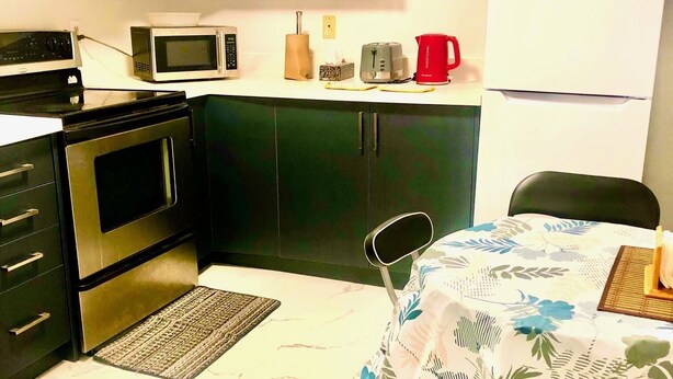 A compact kitchen with a microwave, toaster, and coffee maker on the counter, a refrigerator, and a dining area with a table and chair. - Cozy Home (Markham)