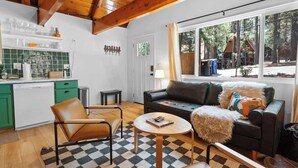 A cozy living room with a sofa, armchair, and coffee table, a kitchen with green cabinets, and a large window with a view of the outdoors.