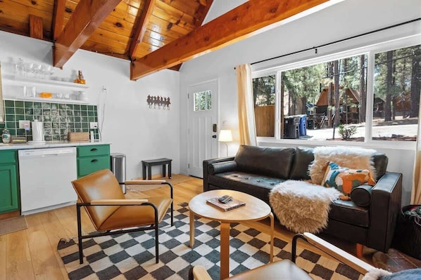 A cozy living room with a sofa, armchair, and coffee table, a kitchen with green cabinets, and a large window with a view of the outdoors.